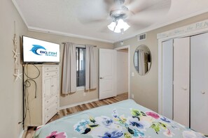 Ein Schlafzimmer mit einem Bett, einem Deckenventilator, einem Fernseher, einem Schminktisch, einem Fenster mit Vorhängen, einem Spiegel und einer Tür.