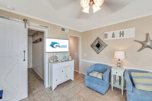 A room with a white door, a flat-screen TV on a white dresser, a blue armchair, a small side table with a lamp, and a wall with a starfish decoration and the word 'BEACH' above it. - Windy Hill Oceanfront Suite, Pool & Hot Tub (North Myrtle Beach)