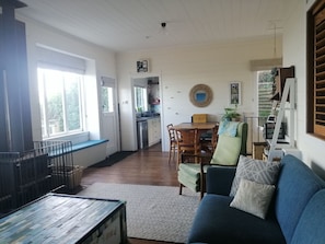 A living room with a blue sofa, wooden dining table, and chairs. There is a kitchen in the background with a window and a plant.