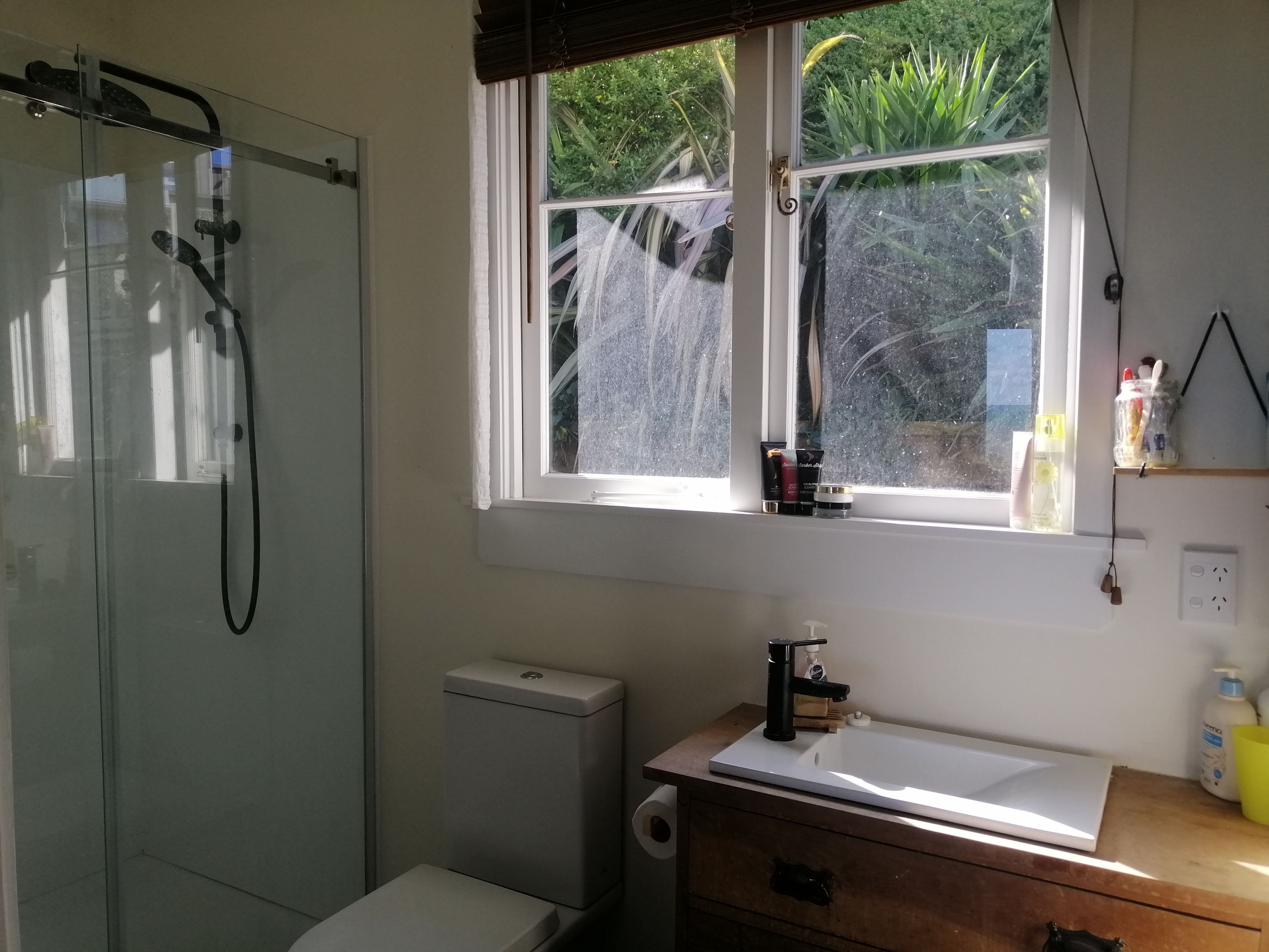 Une salle de bain dotée d’une douche à parois de verre, d’un lavabo blanc, d’un meuble-lavabo en bois et d’une fenêtre donnant sur un paysage verdoyant.