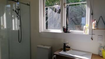 Une salle de bain dotée d’une douche à parois de verre, d’un lavabo blanc, d’un meuble-lavabo en bois et d’une fenêtre donnant sur un paysage verdoyant.