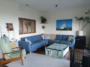 A living room with a blue sofa, a patterned coffee table, a green armchair, a wall-mounted telephone, a painting of a mountain, and a radiator.