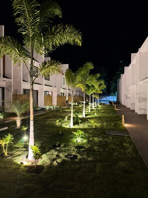 A nighttime view of a modern building with a well-lit garden area featuring young trees and grass. - Beach house in gated community São Miguel dos Milagres CASA MALÚ Alagoas (São Miguel dos Milagres)