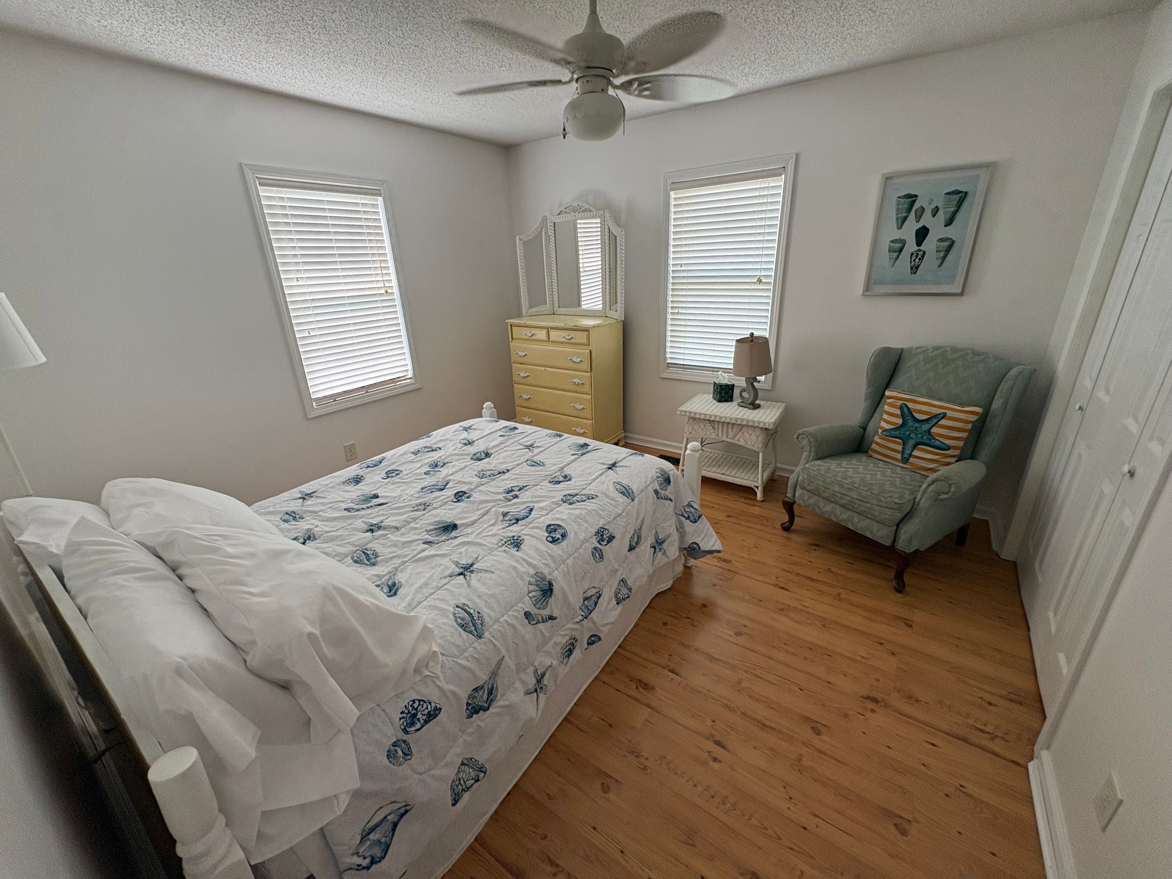 Un dormitorio con cama, cómoda, silla y dos ventanas con persianas.