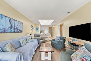 A living room with a sofa, chairs, a coffee table, a TV, and a kitchen in the background. - Pack the Flip‑Flops — Family Fun Starts Here (Atlantic Beach)
