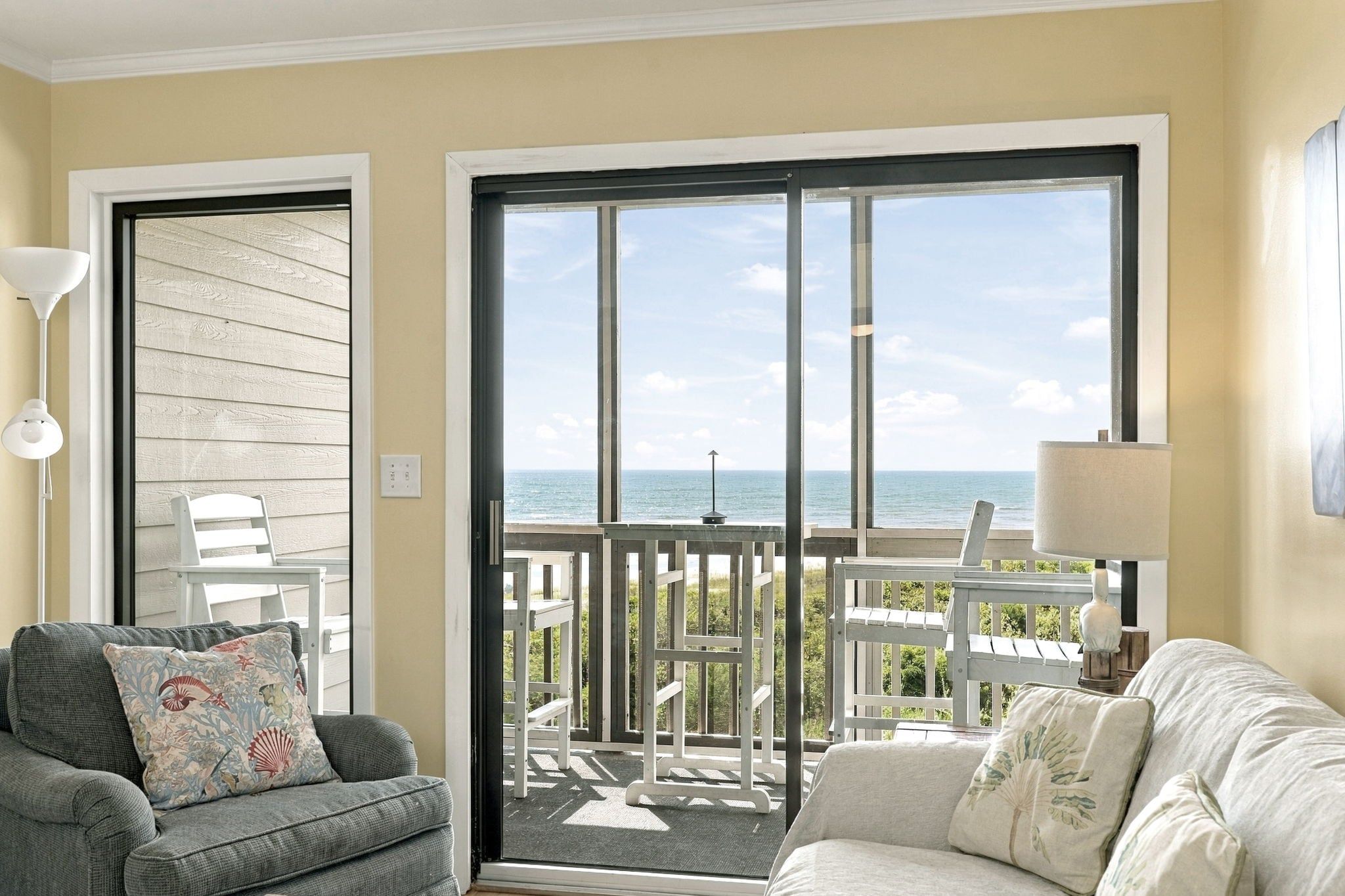 A room with a grey sofa, a white sofa, a table, and a view of the ocean through a sliding glass door.