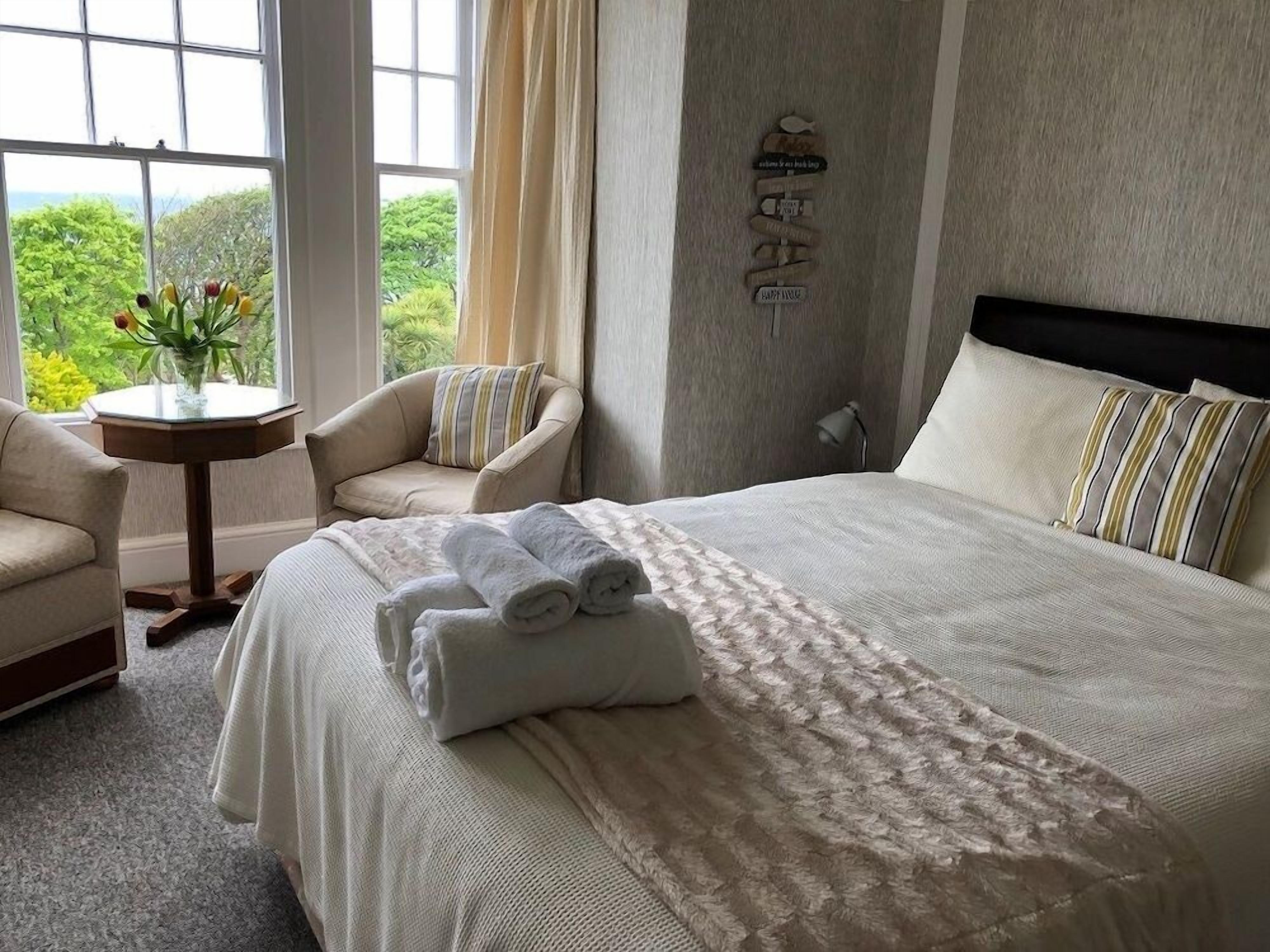 A hotel room with a large bed, a chair, a small table with a vase of flowers, and a view of greenery outside.