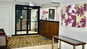 Un hall d’hôtel comprenant un comptoir d’accueil, une entrée avec une porte en verre, un tapis à motifs et un mur orné d’une œuvre d’art représentant un palmier.