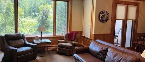A living room with a leather sofa, armchairs, a small table with a lamp, and large windows offering a view of greenery.