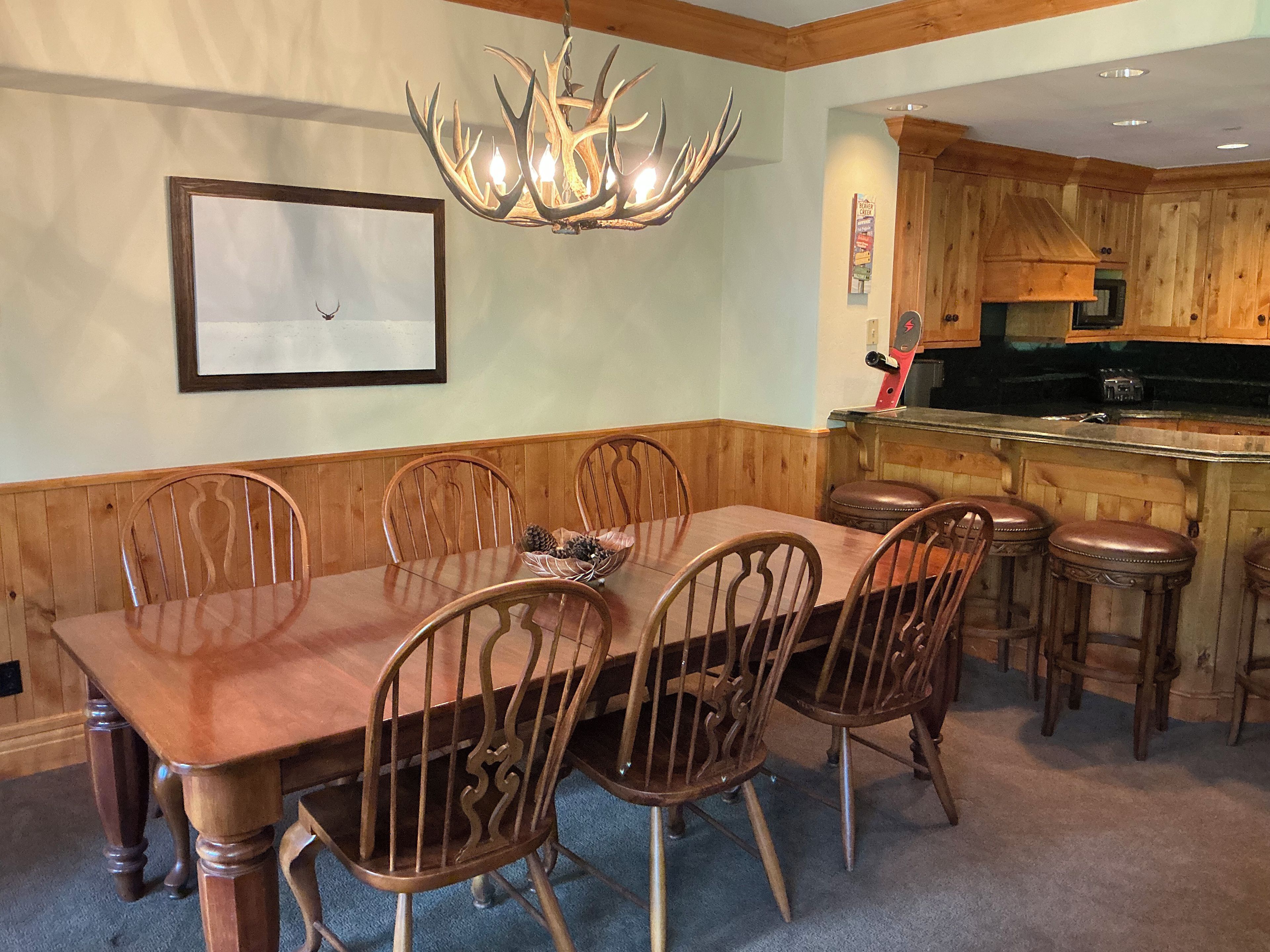 A dining area with a wooden table and chairs, a chandelier, and a kitchen area with wooden cabinets and a bar.