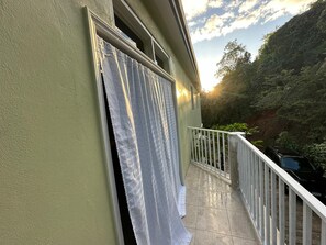 A balcony with a white railing, a window with a curtain, and a view of a forested hillside. - Sun-lit Arched Styled Villa with Scenic Mountain Views and Cool Tranquil Breeze (charlotte amalie)