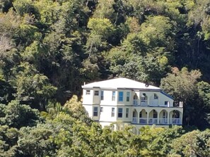 A two-story building with a white facade, multiple windows, and a balcony. It is surrounded by dense greenery. - Sun-lit Arched Styled Villa with Scenic Mountain Views and Cool Tranquil Breeze (charlotte amalie)