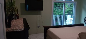 A hotel room with a bed, a TV mounted on the wall, a window with a view of greenery, and a balcony with a chair. - Sun-lit Arched Styled Villa with Scenic Mountain Views and Cool Tranquil Breeze (charlotte amalie)