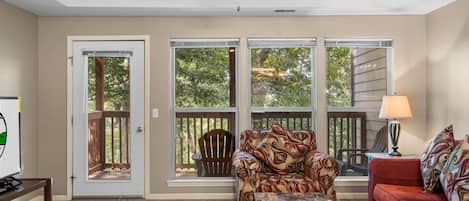 A living room with a ceiling fan, a red couch, a patterned armchair, a glass coffee table, and a view of trees through large windows.