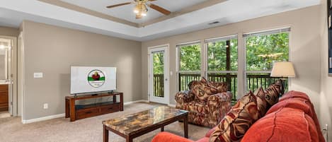 A living room with a ceiling fan, a television on a wooden stand, a patterned sofa, and a window with a view of trees.