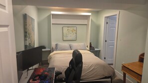 Ein kleines, helles Schlafzimmer mit einem Bett, einem Schreibtisch mit Computer und Kopfhörern, einem Stuhl und einer Tür.