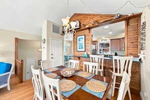 A dining area with a wooden table and white chairs, a kitchen with wooden cabinets, and a view of the beach. - Beach Bungalow: Soundside, Hot Tub, Sound Views, Village Beach Club Access (Nags Head)