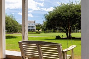 A porch with a swing chair overlooking a well-maintained lawn and a two-story house with a balcony. - Beach Bungalow: Soundside, Hot Tub, Sound Views, Village Beach Club Access (Nags Head)