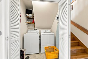 A laundry room with two washing machines, a dryer, a yellow laundry basket, and a shelf with a red item. - Beach Bungalow: Soundside, Hot Tub, Sound Views, Village Beach Club Access (Nags Head)