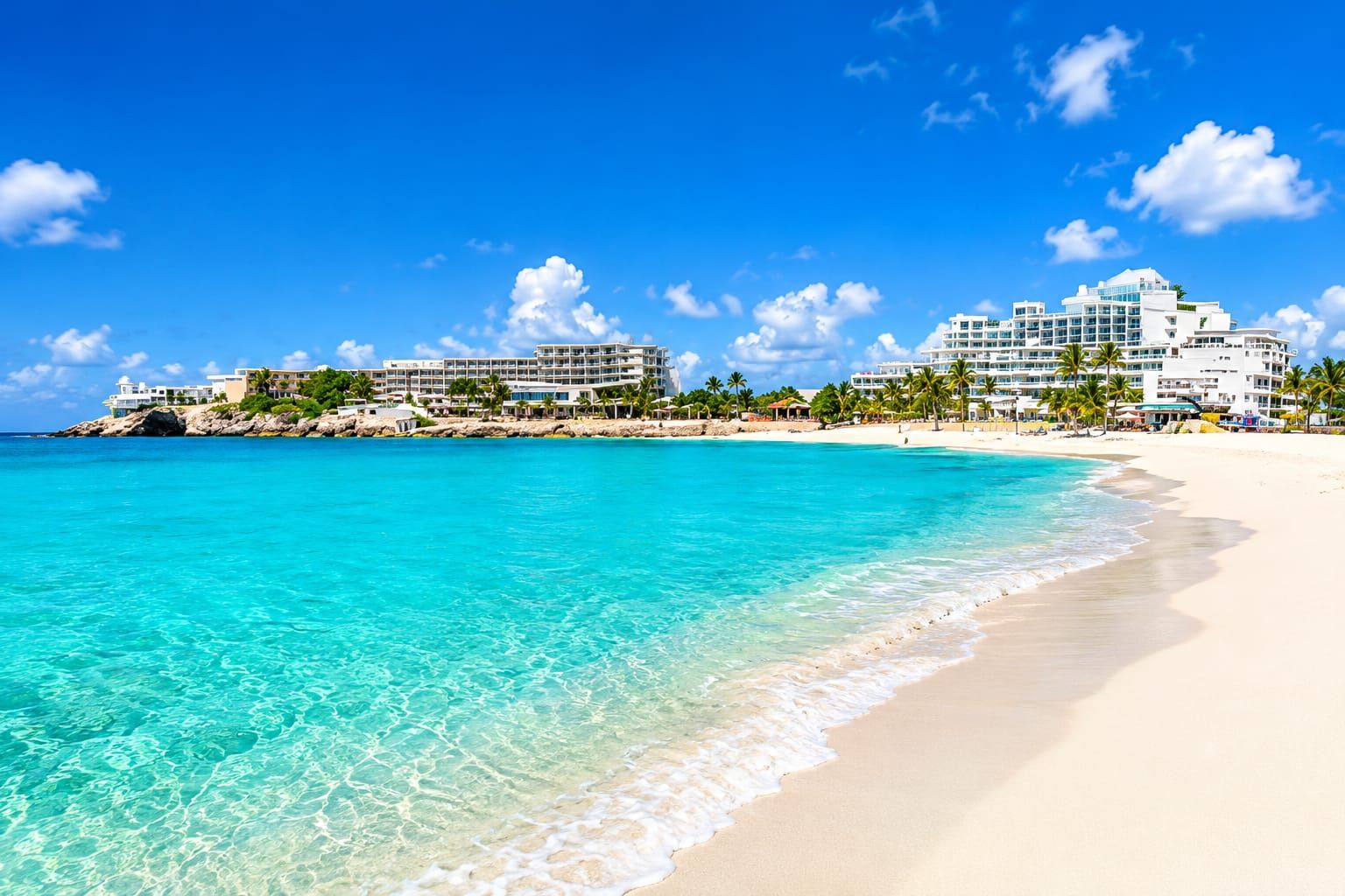 A beach with clear turquoise water, white sand, and a resort with multiple buildings and palm trees.