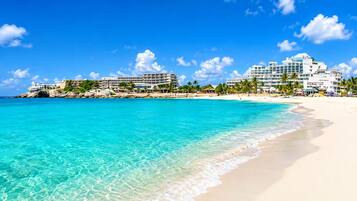 A beach with clear turquoise water, white sand, and a resort with multiple buildings and palm trees.
