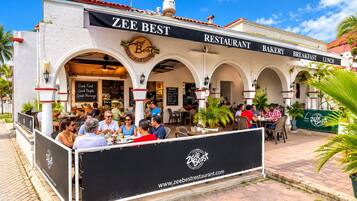 A restaurant with outdoor seating, a sign reading 'Zee Best Restaurant Bakery Breakfast Lunch,' and a website address 'www.zeebestrestaurant.com' on a banner.