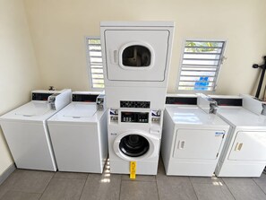 Ein Waschraum mit vier Waschmaschinen und Trocknern, einem Bedienfeld und Fenstern mit Jalousien.