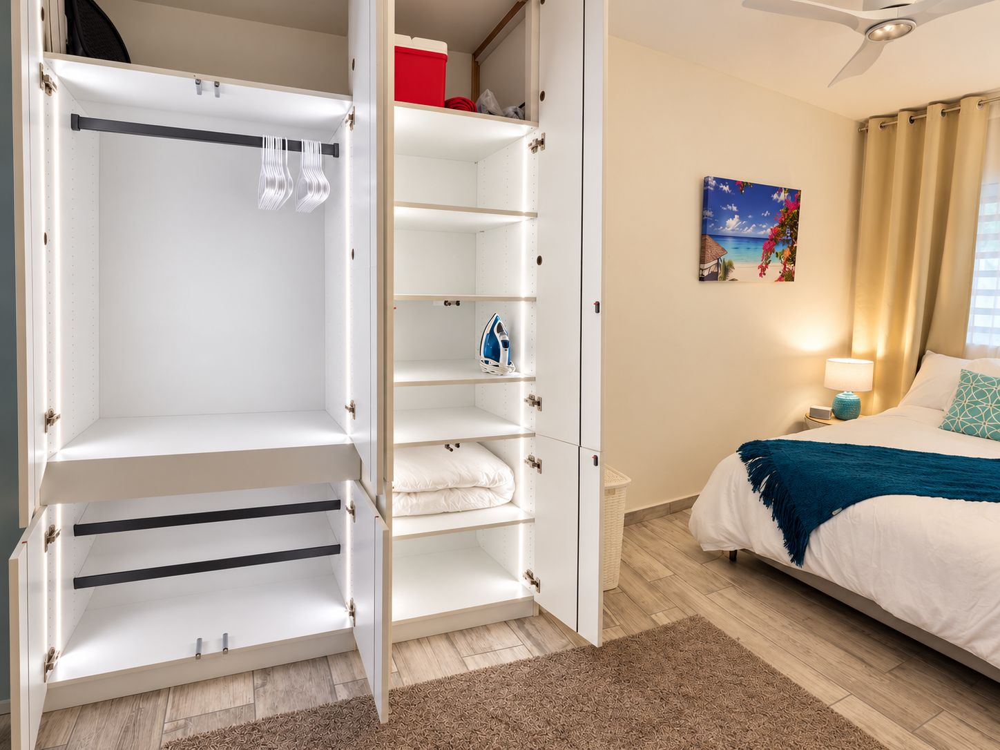 A hotel room with a wardrobe, a bed, a nightstand, a lamp, and a painting on the wall.