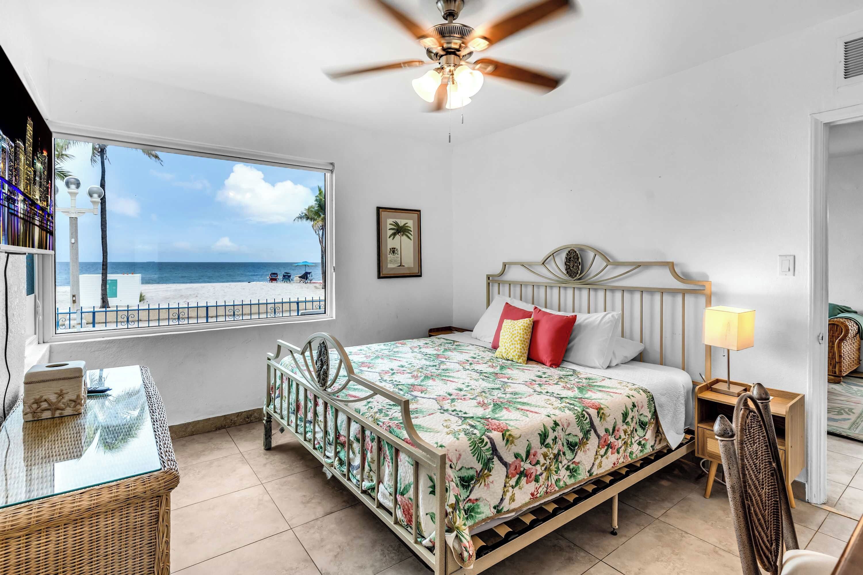 A bedroom with a bed, a ceiling fan, a window with a view of the beach, and a bedside table with a lamp.