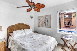 Un dormitorio con una cama, un ventilador de techo, una lámpara de pared, una ventana con vistas y un cuadro de loros.