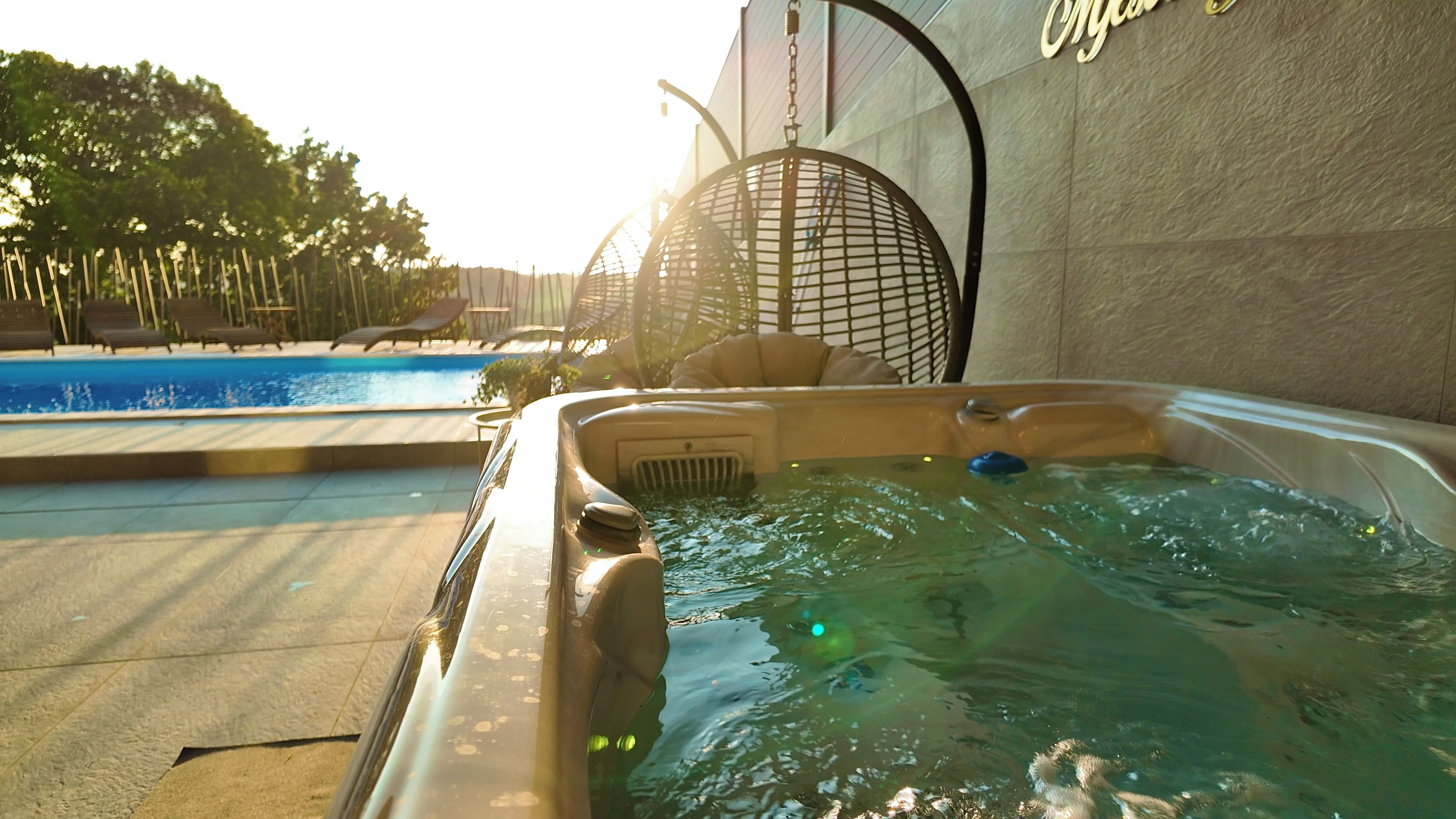 A hot tub with a pool in the background, surrounded by lounge chairs and a pergola.