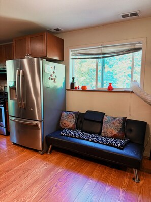 A compact kitchen with a stainless steel refrigerator, a black sofa with patterned cushions, and a window with a view of trees.