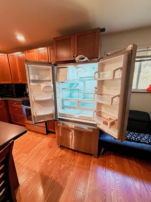 A stainless steel refrigerator with a freezer compartment, situated in a kitchen with wooden cabinets and a window. - Cross Creek Cabin - Private Redwoods, 2 Creeks, Massage Bed & 100" Theater (Redway)