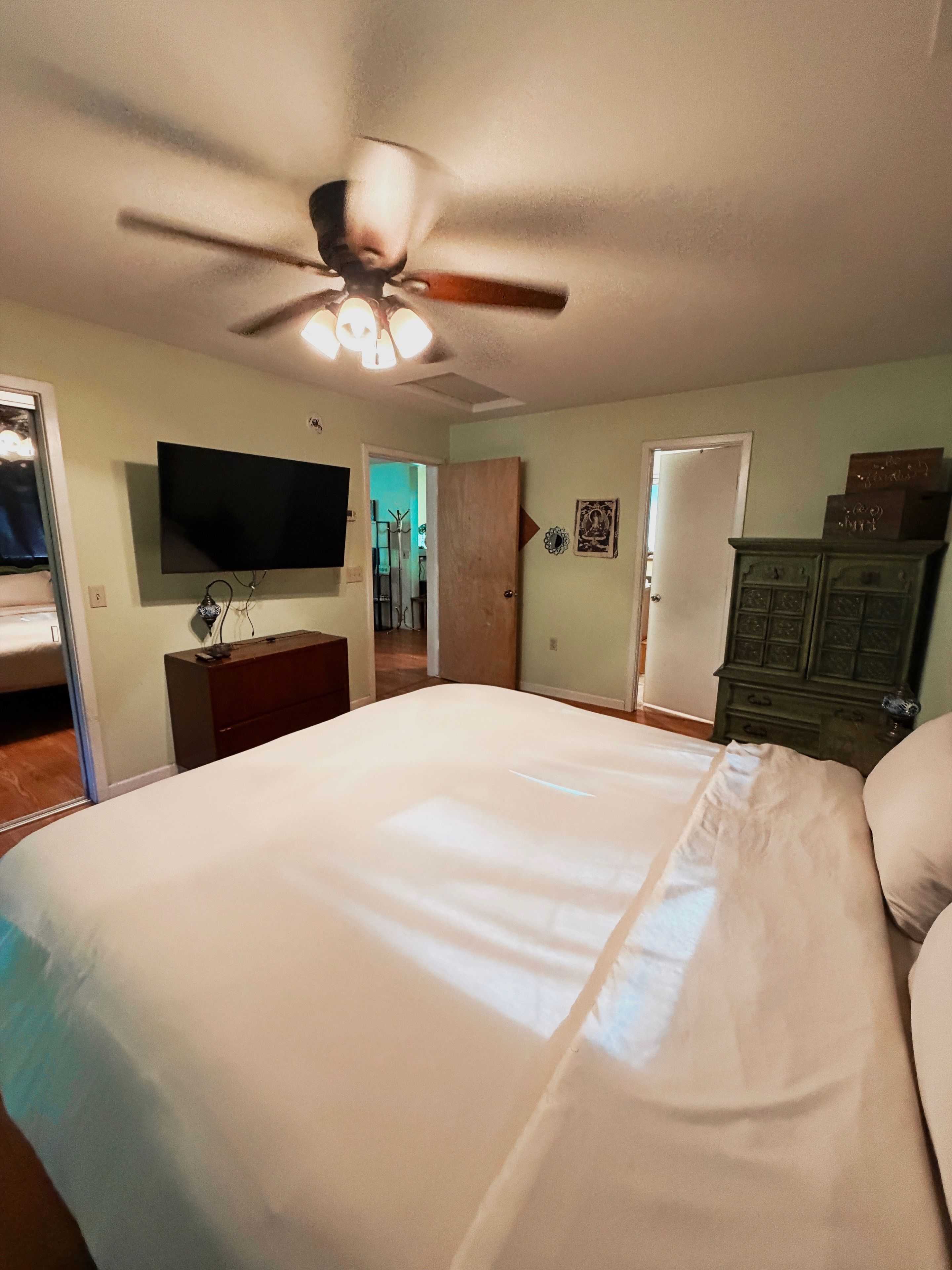 Ein Schlafzimmer mit Bett, Deckenventilator, Fernseher, Kommode und einer Tür.