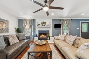 A living room with a fireplace, a ceiling fan, and a television mounted on the wall. - Waco Woodland Retreat, Sleeps 14! Waco/Baylor (Waco)