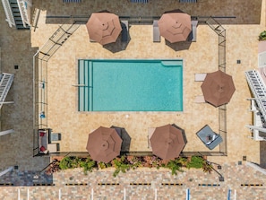 An aerial view of a rooftop pool area with a rectangular pool, four umbrellas, and a seating area with chairs and a sofa. - Poolside Fun, Pickleball & Coastal Adventures Await in Fort Myers - Jefferson Palms I - Roelens (Fort Myers Beach)