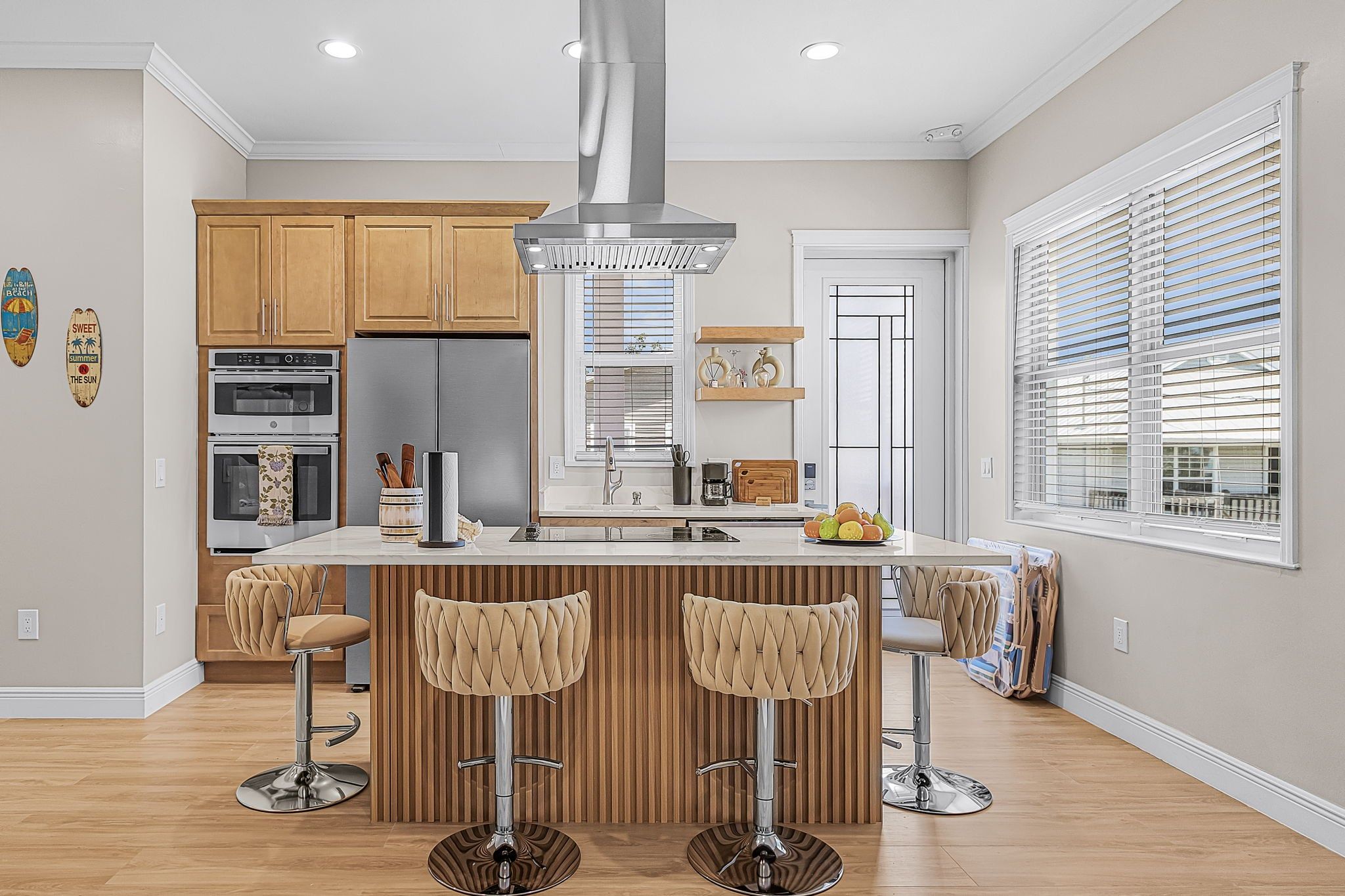 A modern kitchen with wooden cabinets, stainless steel appliances, a central island with bar stools, and a large window with blinds.