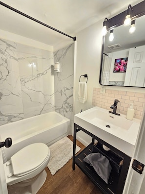 Un baño con inodoro blanco, bañera, lavamanos con espejo y toallero.