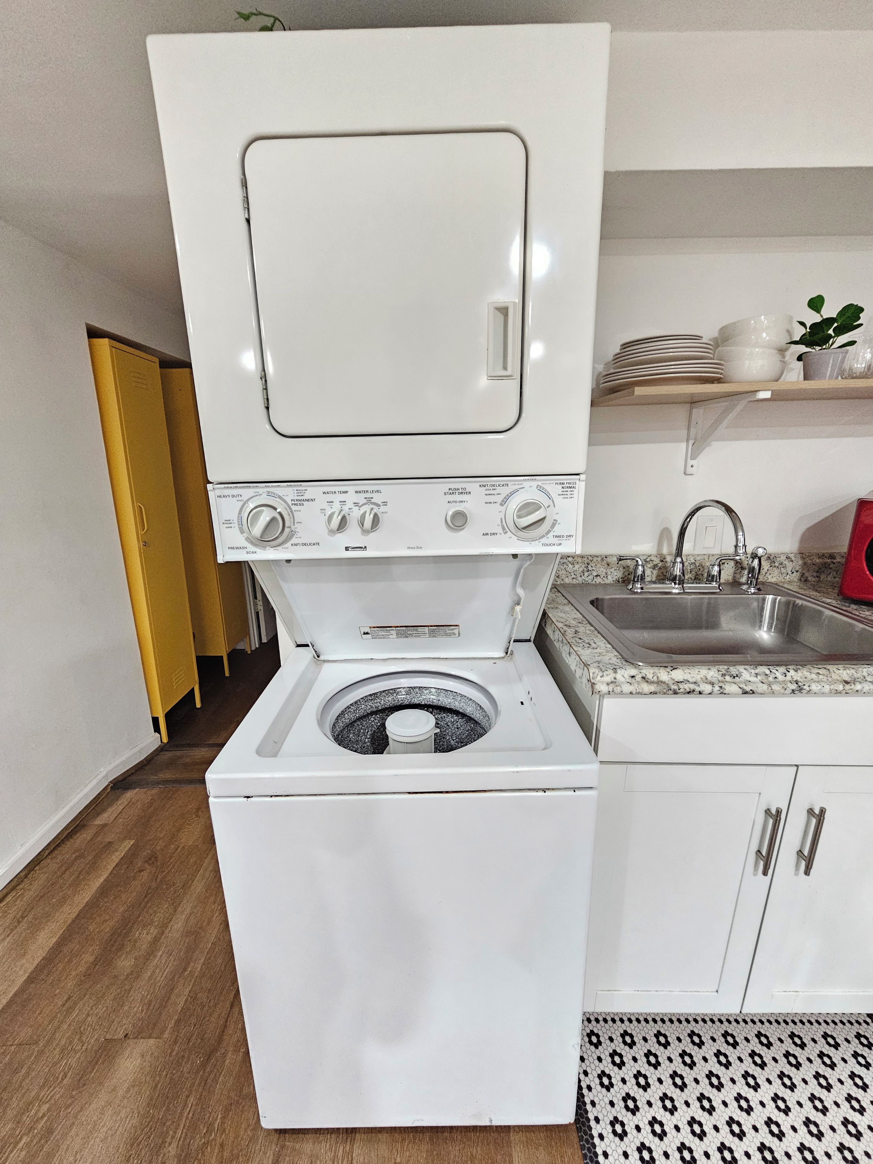 A compact, white washing machine with a front-loading door, situated in a small kitchen area with a granite countertop and a stainless steel sink.