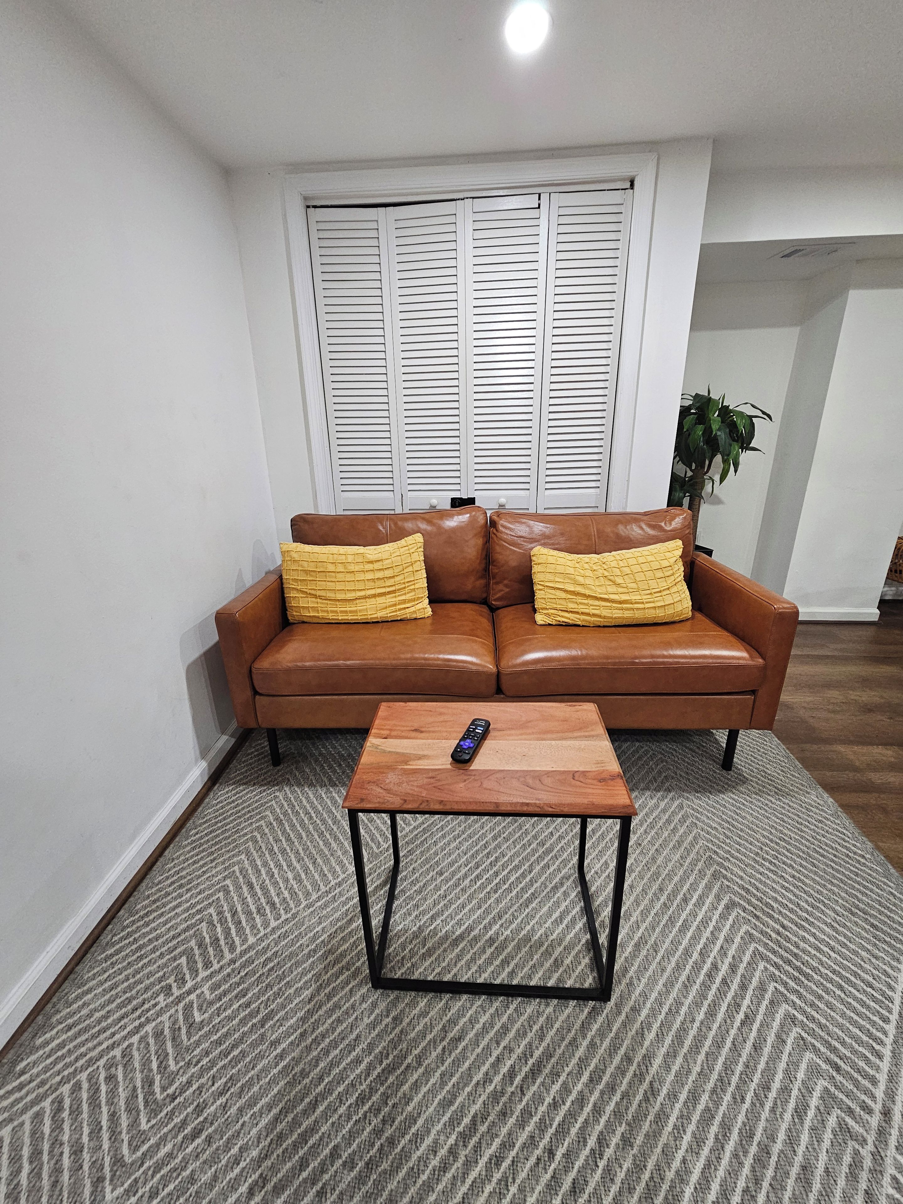 A living room with a brown leather sofa, two yellow cushions, a wooden coffee table, and a remote control on it.