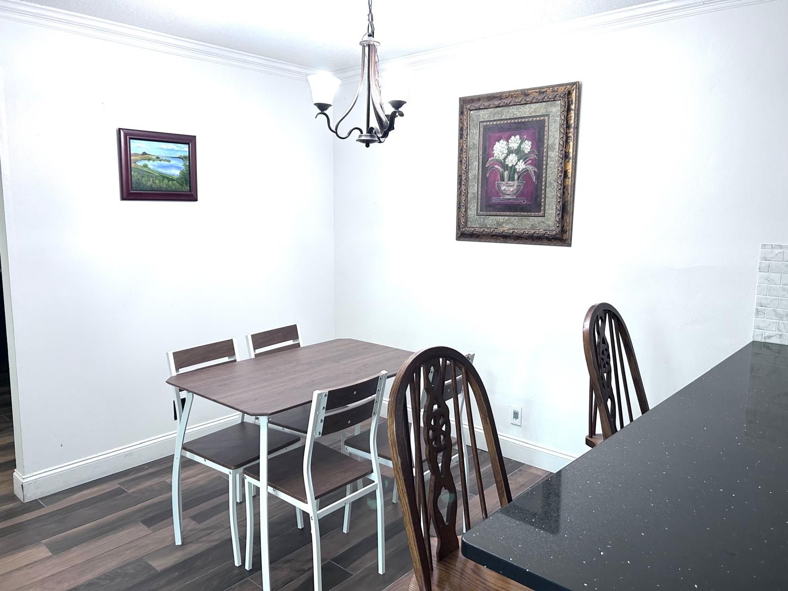 Een eetgedeelte met een rechthoekige tafel, vier stoelen, een kroonluchter en twee ingelijste foto's aan de wanden.
