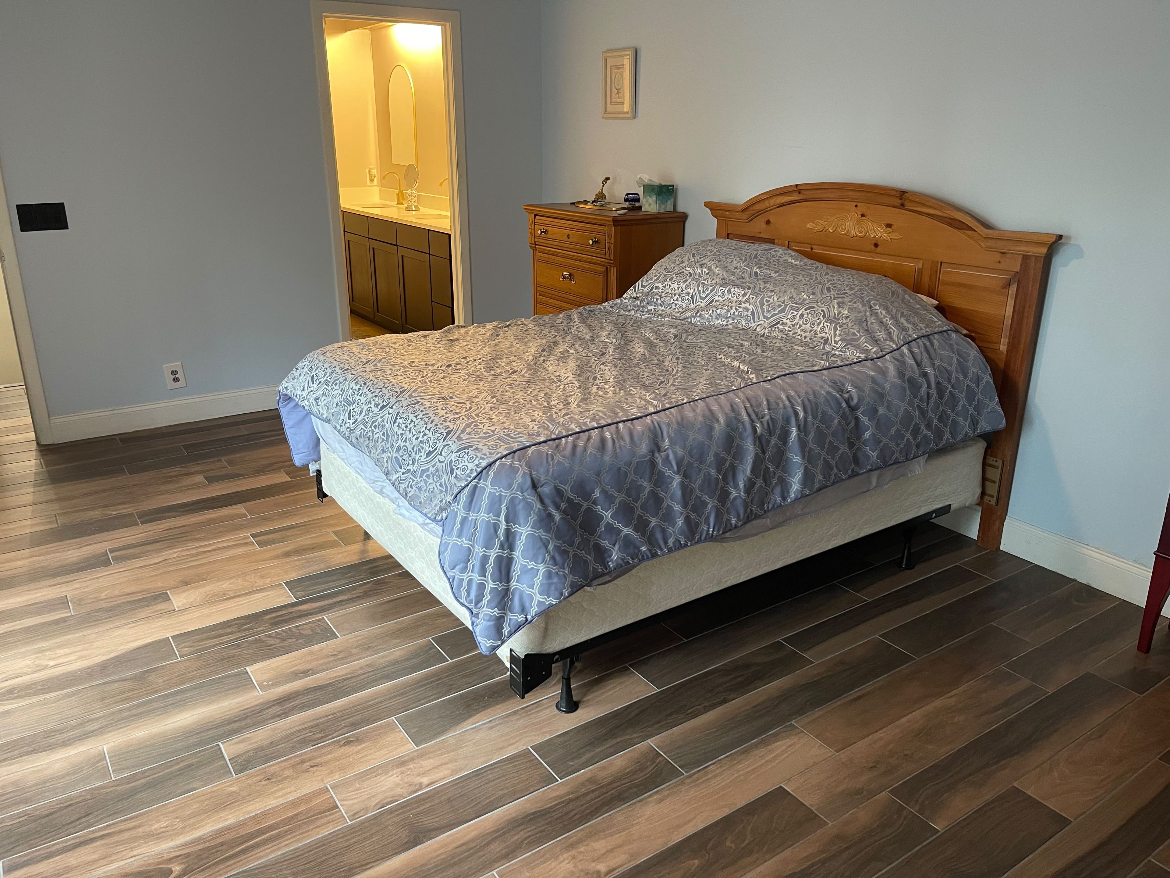 A bedroom with a bed, wooden headboard, nightstand, and a bathroom with a mirror and sink.