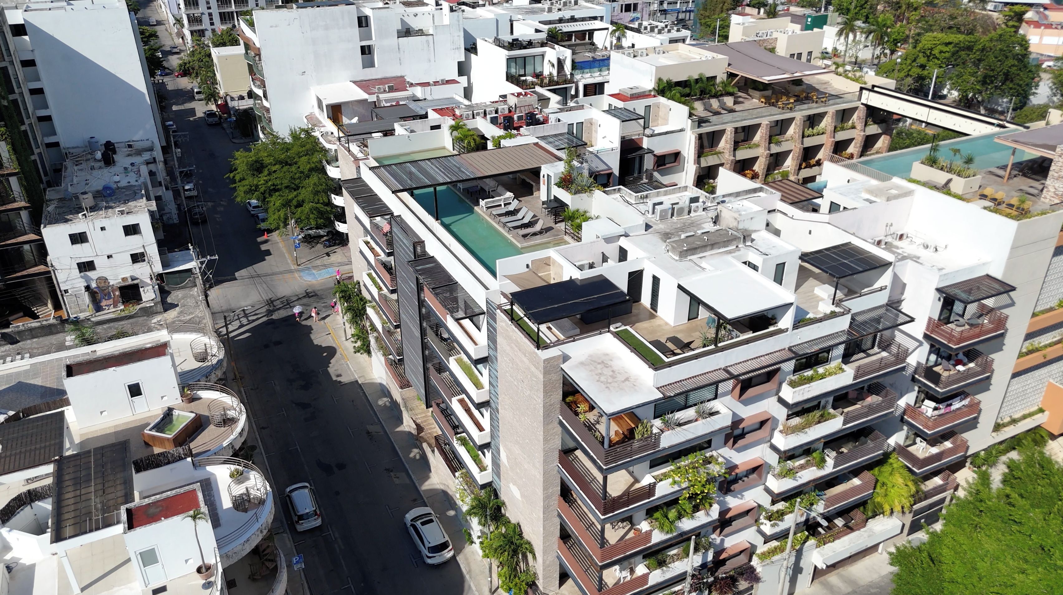 A modern urban residential area with multi-story buildings, a central road, and a swimming pool on one of the rooftops.