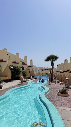 A swimming pool with a wavy edge, surrounded by beige buildings with red accents, and palm trees. - Mareverde Sea View Apartment with Pool (Costa Adeje)