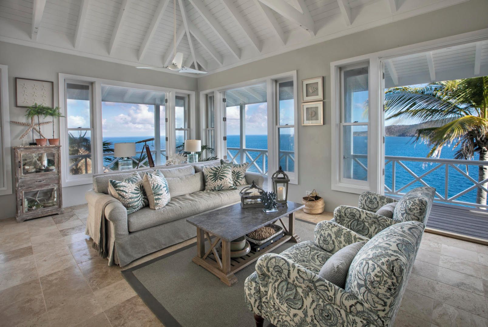 A spacious living room with a large sofa, patterned armchairs, and a coffee table. There are multiple windows offering ocean views.