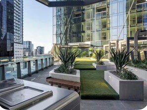 A modern rooftop terrace with a glass building, planters, and a pathway. - Modern CBD Retreat | Pool, Sauna, Gym, BBQ  & City Views (Melbourne)