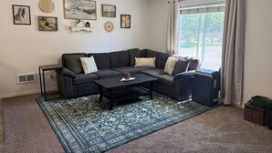 A living room with a sectional sofa, a coffee table, and a large window with curtains. - The Travelers Nest | BYUI, Island Park, & Targhee (Rexburg)