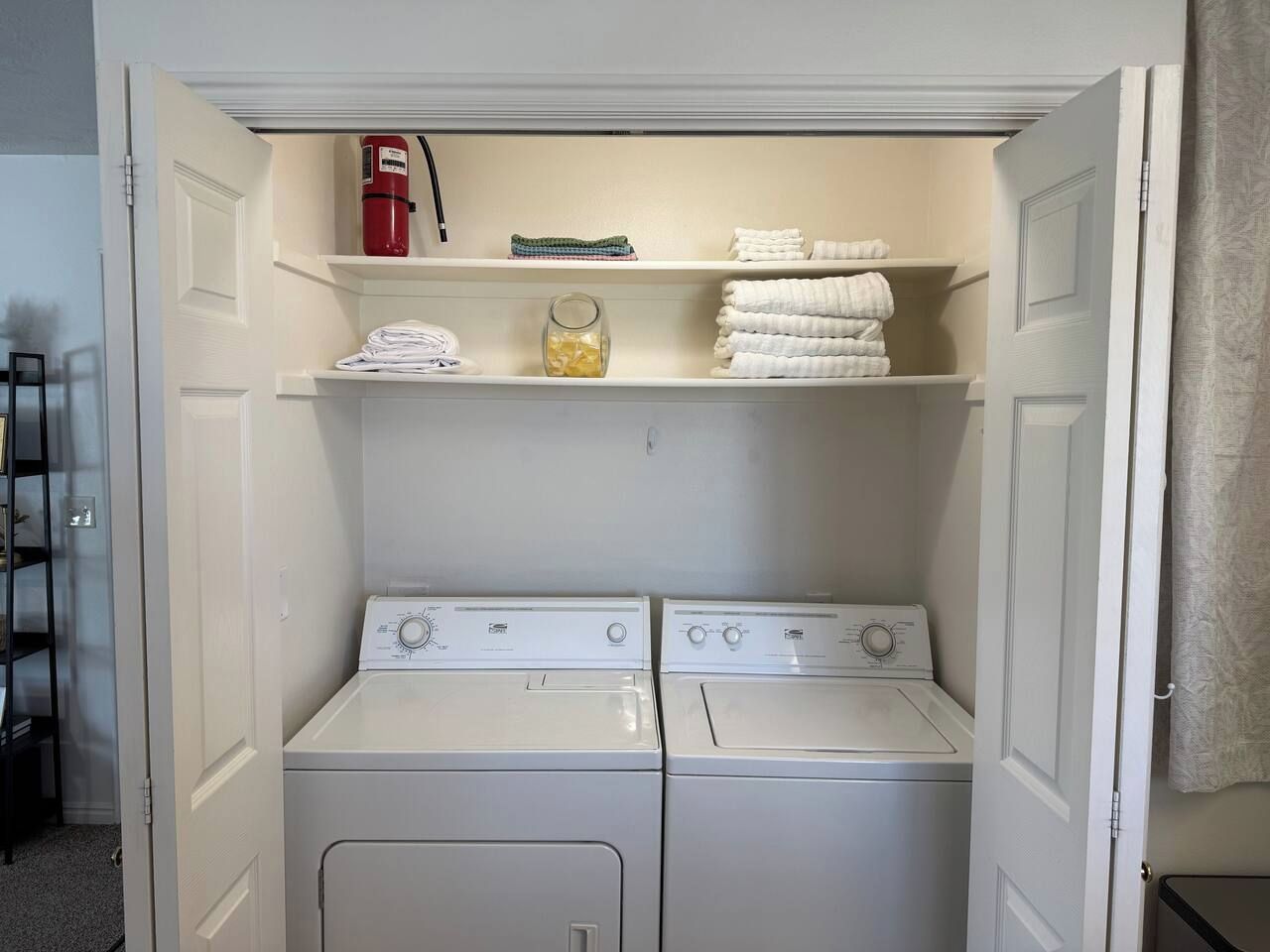 A laundry room with two stacked washers and dryers, shelves with folded towels, and a fire extinguisher.