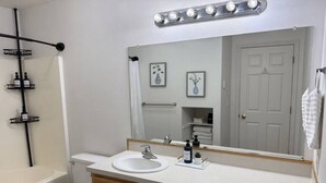 A bathroom with a white sink, wooden vanity, a mirror with lights, a toilet, and a shower area. - The Travelers Nest | BYUI, Island Park, & Targhee (Rexburg)