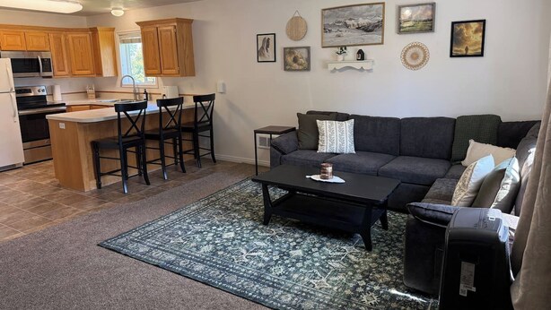 A living room with a sofa, coffee table, and a rug. There is a kitchen area with wooden cabinets and a microwave. - The Travelers Nest | BYUI, Island Park, & Targhee (Rexburg)